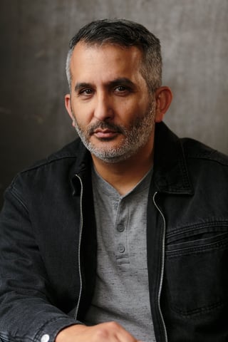 Portrait of a middle-aged man with gray-streaked dark hair and beard wearing a black jacket over a gray shirt against a neutral background