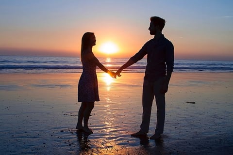 Couple holding hands on beach at sunset, silhouetted against golden sky and ocean waves