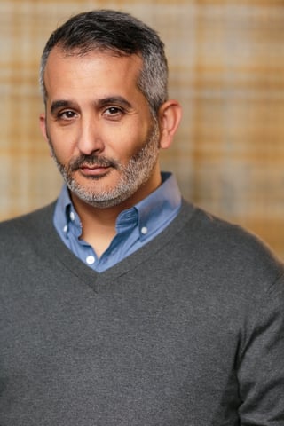 Professional headshot of a middle-aged man with gray hair and beard wearing a gray sweater over a blue shirt against a blurred beige background