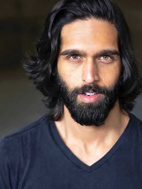 Man with long black hair and full beard wearing a dark v-neck shirt, smiling at camera against blurred background