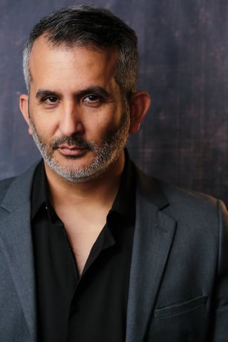 Portrait of a middle-aged man with short dark hair and beard wearing a dark blazer and black shirt against a blurred grey background