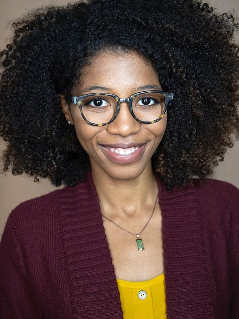 Portrait of a smiling woman with curly dark hair, wearing glasses, a burgundy cardigan, and a yellow top with a green pendant necklace