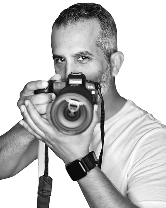 A man with a beard wearing a white shirt holds a professional DSLR camera up to his face, aiming directly at the viewer in a black and white portrait