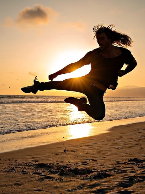 Silhouette of a person performing a jumping kick on a beach at sunset with golden light and ocean waves