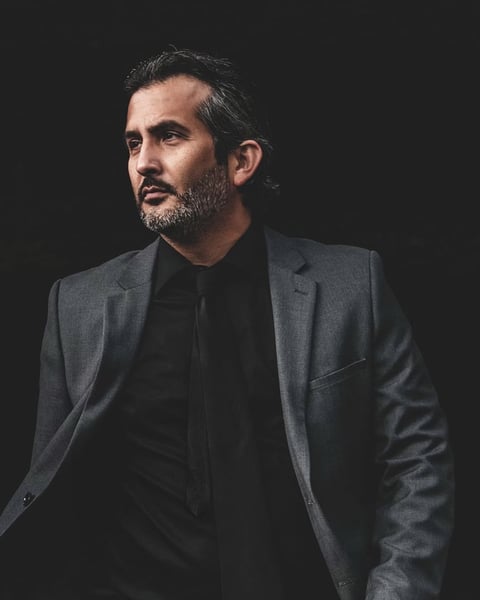 Man with dark hair and beard wearing a gray blazer over black shirt
