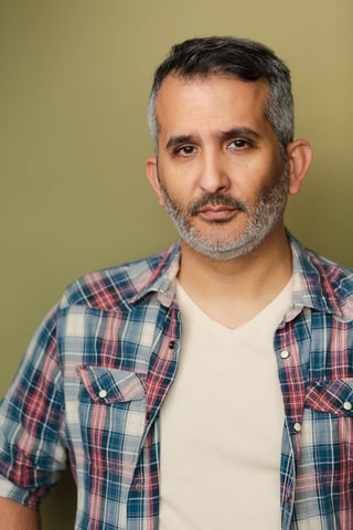 Middle-aged man with gray beard wearing a blue and red plaid shirt over a white v-neck shirt against a pale green background