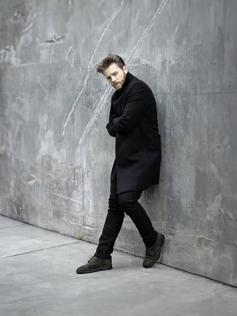 Man in black coat and boots leaning against weathered gray concrete wall