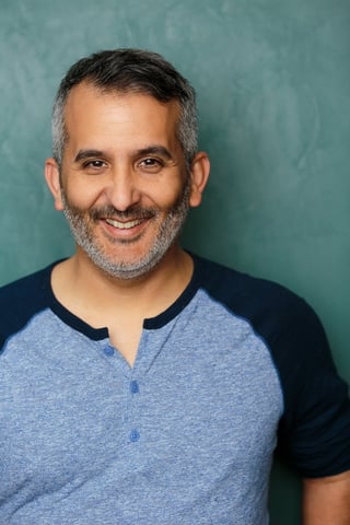 Smiling man with gray beard wearing a blue and black baseball tee against a teal background