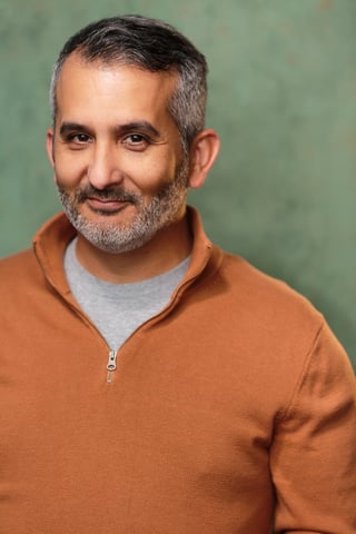 Portrait of a middle-aged man with gray beard wearing an orange quarter-zip sweater over a gray shirt against a green background