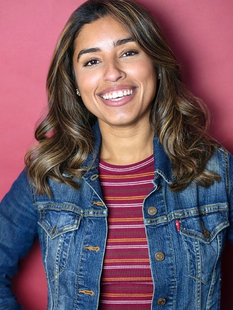 Young woman smiling at camera wearing a denim jacket and red striped shirt against a pink background
