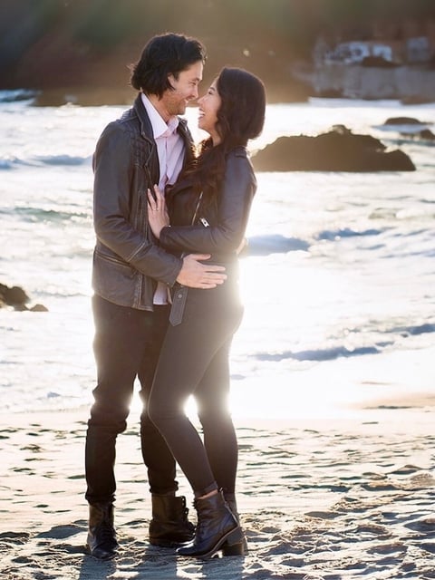 Couple embracing on beach at sunset with waves and rocky shoreline in background