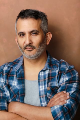 Man with gray beard wearing blue plaid shirt over gray t-shirt, arms crossed, against tan background