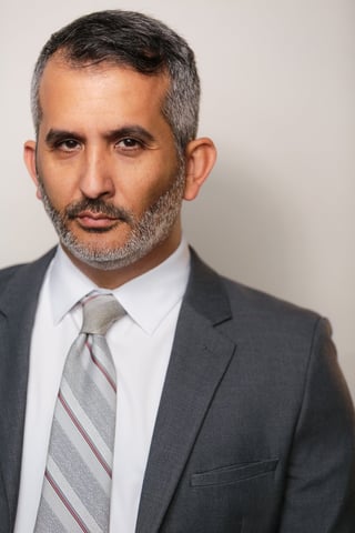 Professional headshot of a man with gray hair and beard wearing a charcoal suit, white shirt, and striped tie against a neutral background